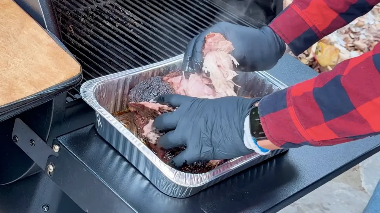 Gloved hands pulling apart cooked pork shoulder in an aluminum pan
