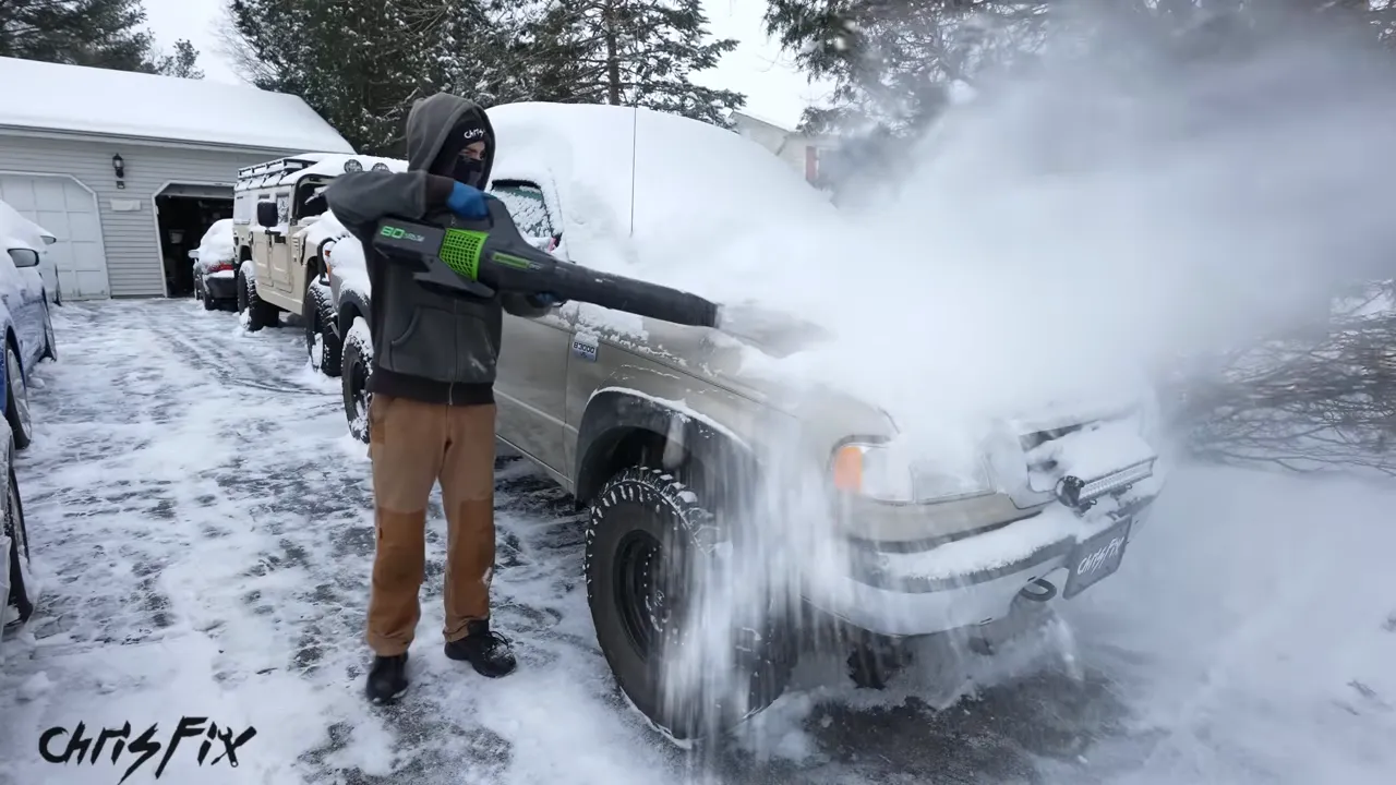 A person using a leaf blower to blow snow off the hood of a pickup truck