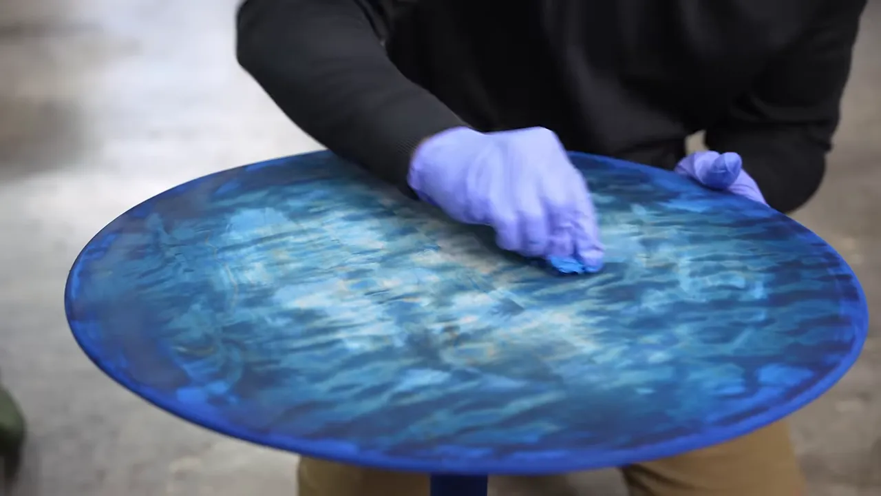 A quilted maple tabletop with a turquoise center and dark blue perimeter being wiped with a cloth