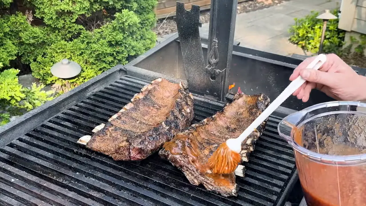 A brush spreading red barbecue glaze over beef rib racks on the grill