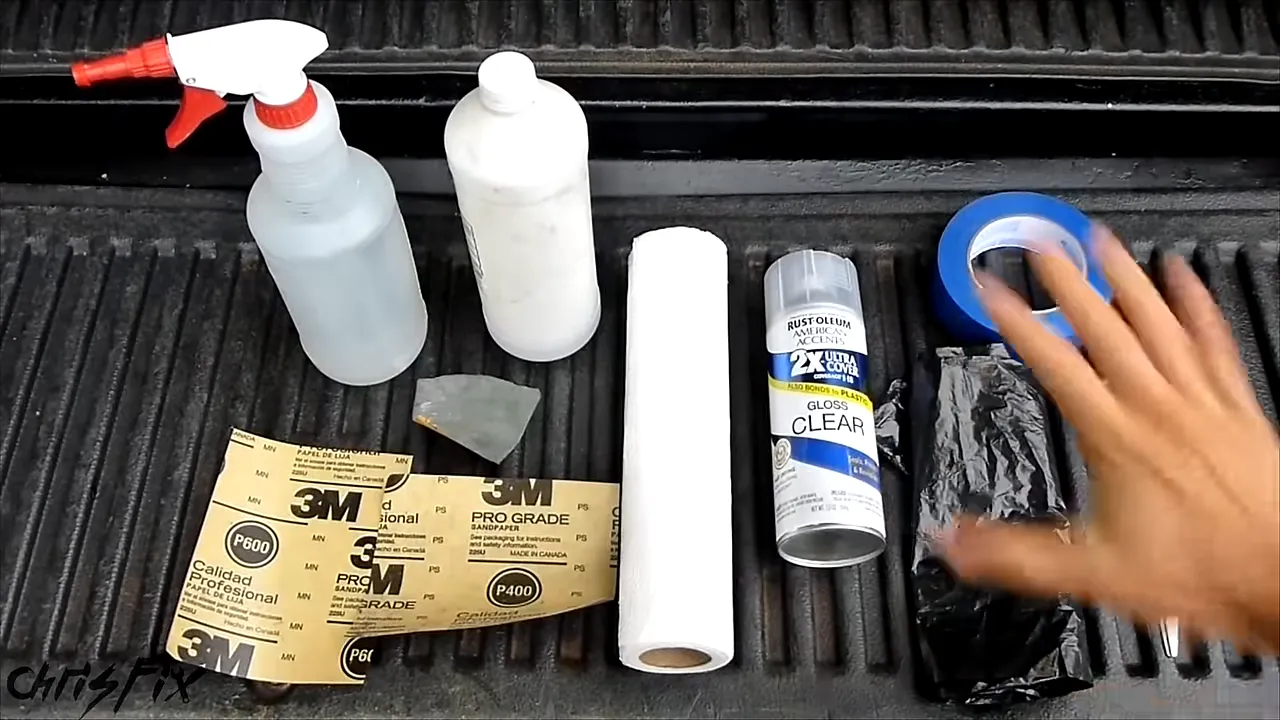 Sandpaper, spray bottle, rubbing alcohol, clear coat, paper towels, tape, garbage bag, and scissors on a truck bed