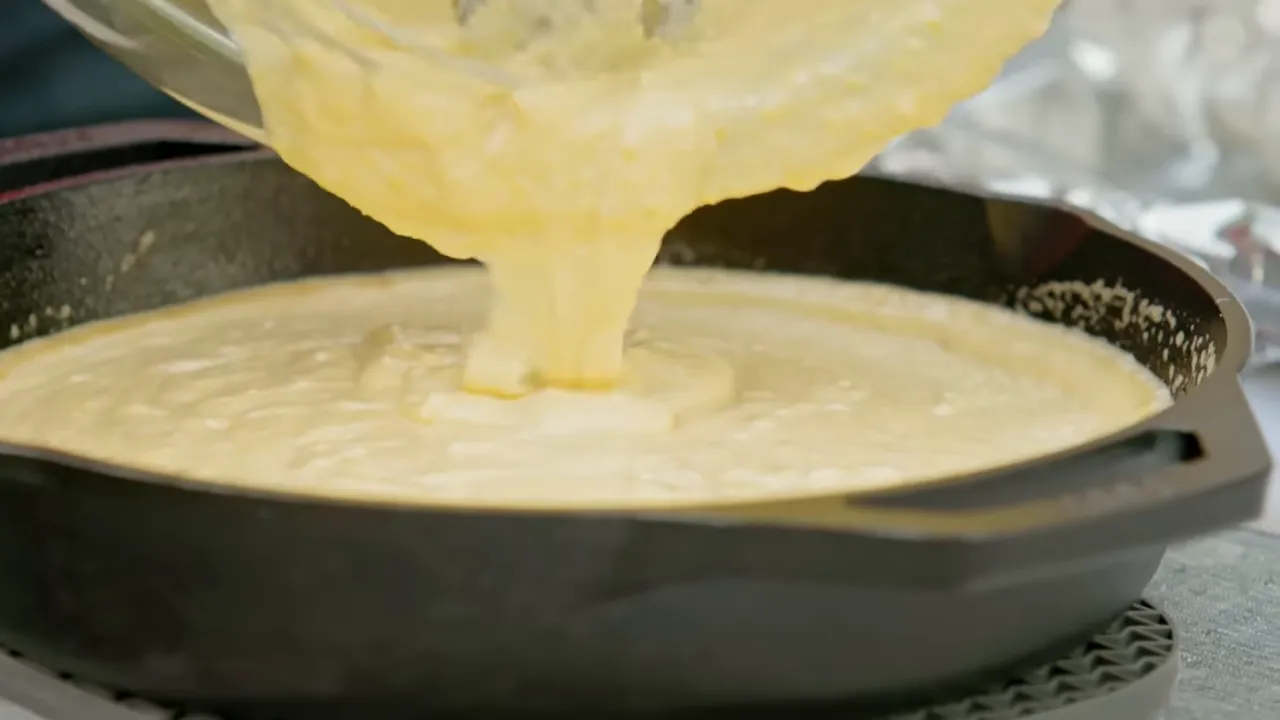 Pale cornbread batter being poured into a buttered cast iron skillet