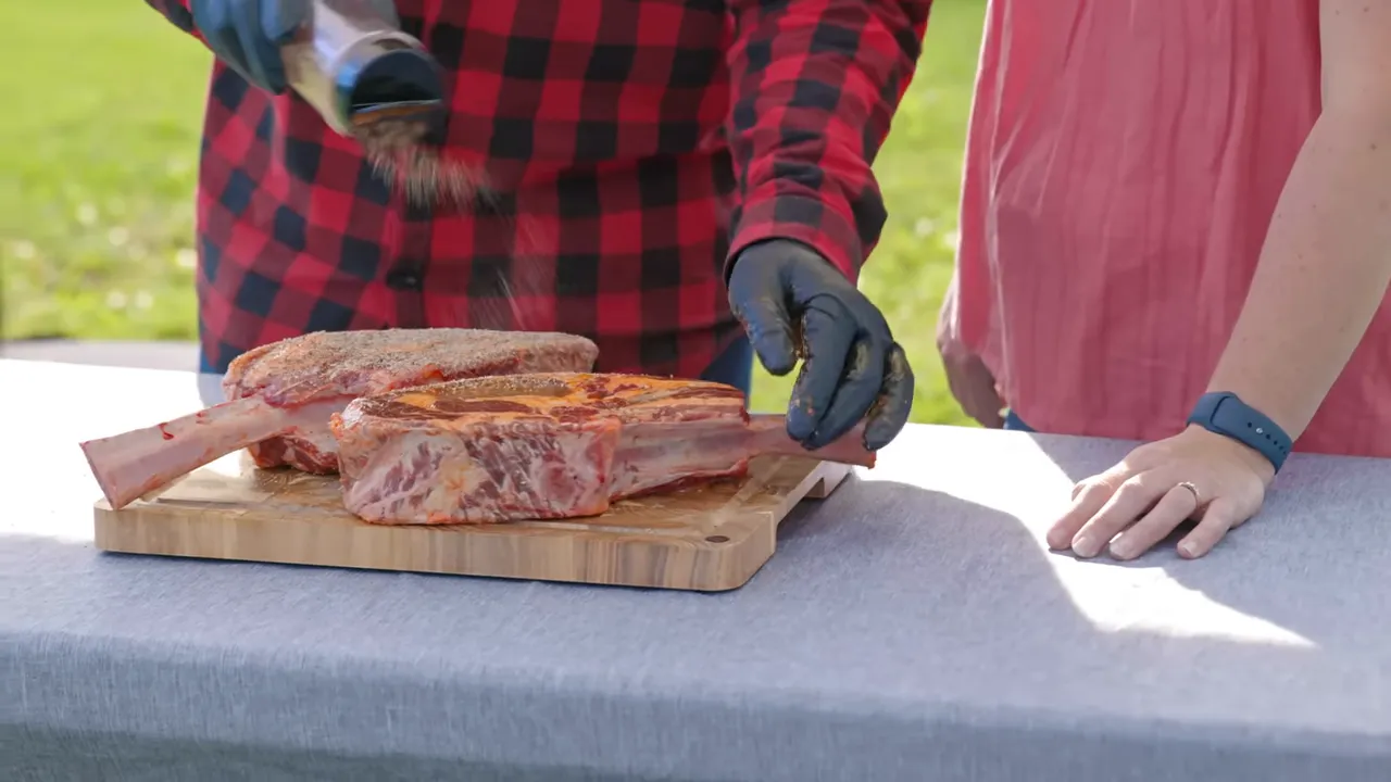 Seasoning being sprinkled over two tomahawk steaks on a wooden board