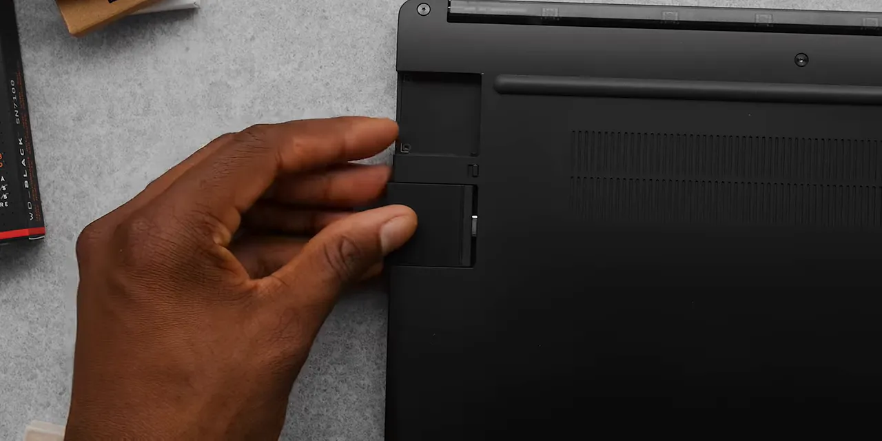 A hand inserting a silver expansion card into the underside of the Framework laptop
