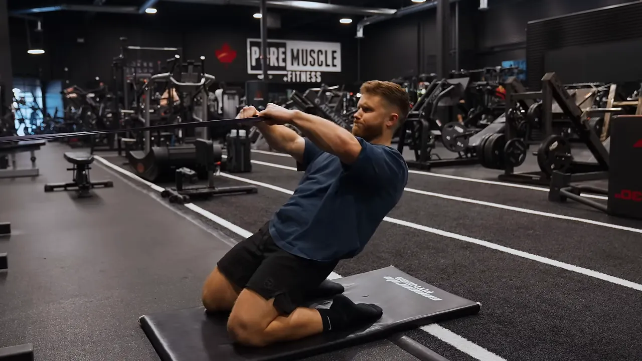 Lifter kneeling on a mat and leaning backward during a reverse Nordic while holding a band