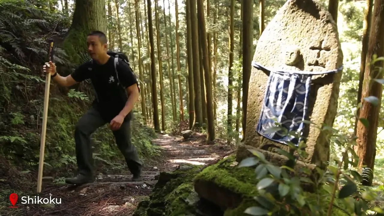 Hiker with a walking staff on a forest pilgrimage trail in Shikoku passing a moss-covered stone Jizo statue.