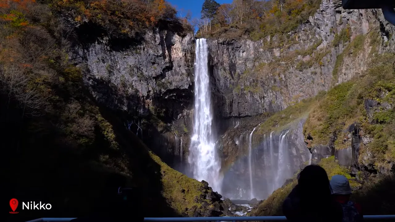 Close-up of Kegon Falls plunging between rocky cliffs with mist at the base, autumn foliage on the rim and silhouetted visitors on a viewing platform