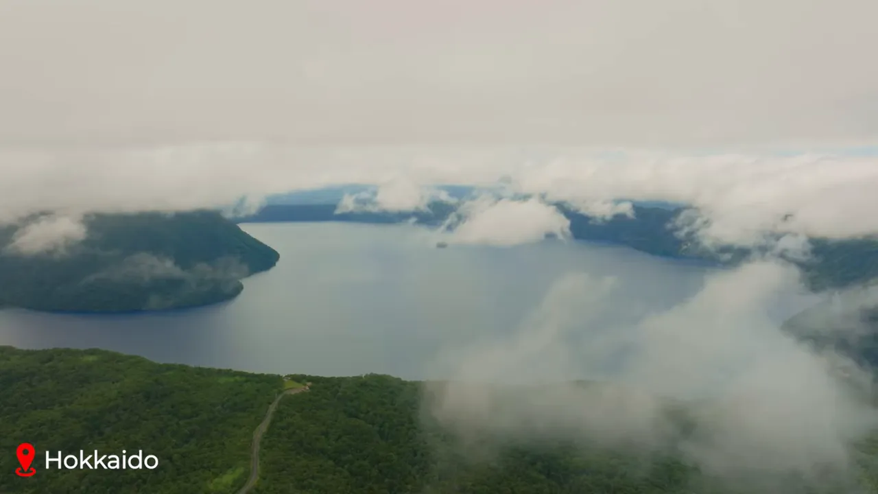 Wide aerial view of a misty lake surrounded by forested hills in Hokkaido with low clouds drifting across the water
