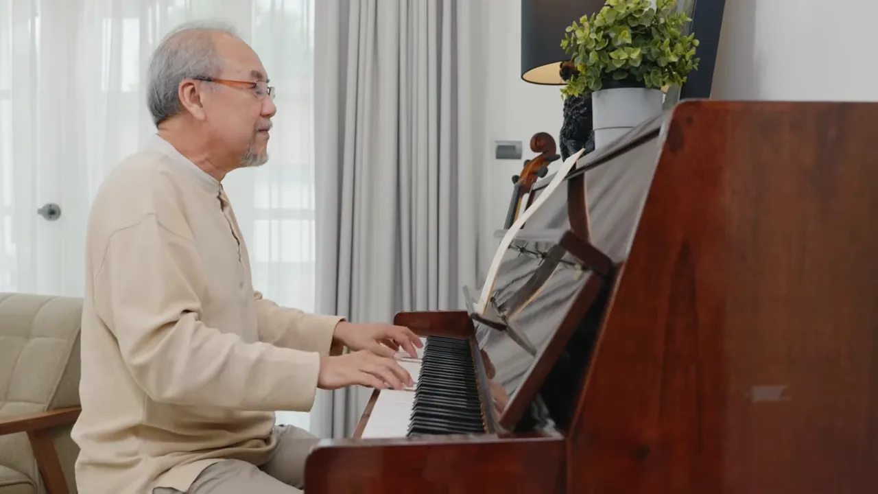 Elderly man seated at an upright piano playing indoors, looking focused and engaged