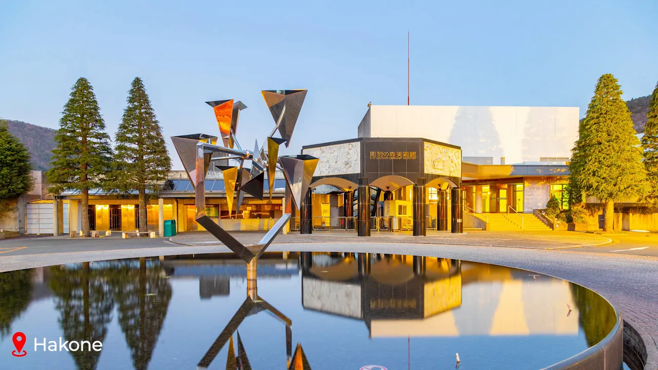 Contemporary outdoor sculpture standing over a reflecting pond with the museum building and trees in the background in Hakone