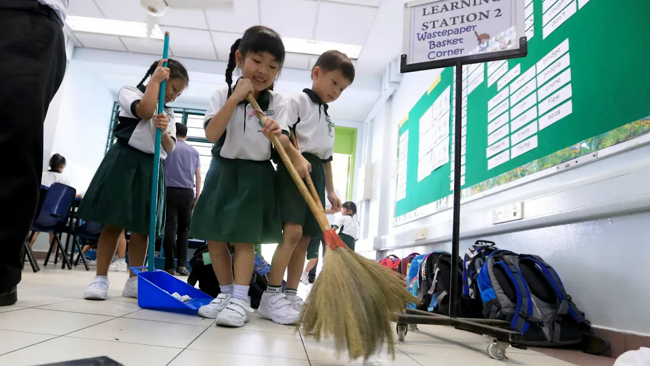 Primary school students in uniform sweeping a classroom floor with brooms and dustpans during daily cleaning time