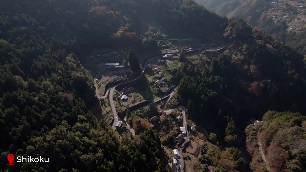 Wide aerial view of a rural Shikoku valley showing clustered houses, winding roads and forested hills — the landscape of the 88-temple pilgrimage