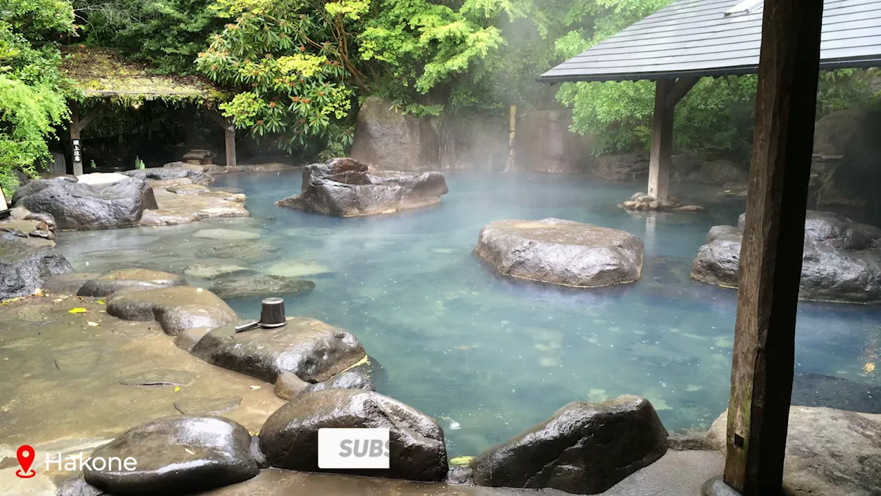 Natural outdoor onsen pool with steaming blue water, large rocks and green trees surrounding the bath