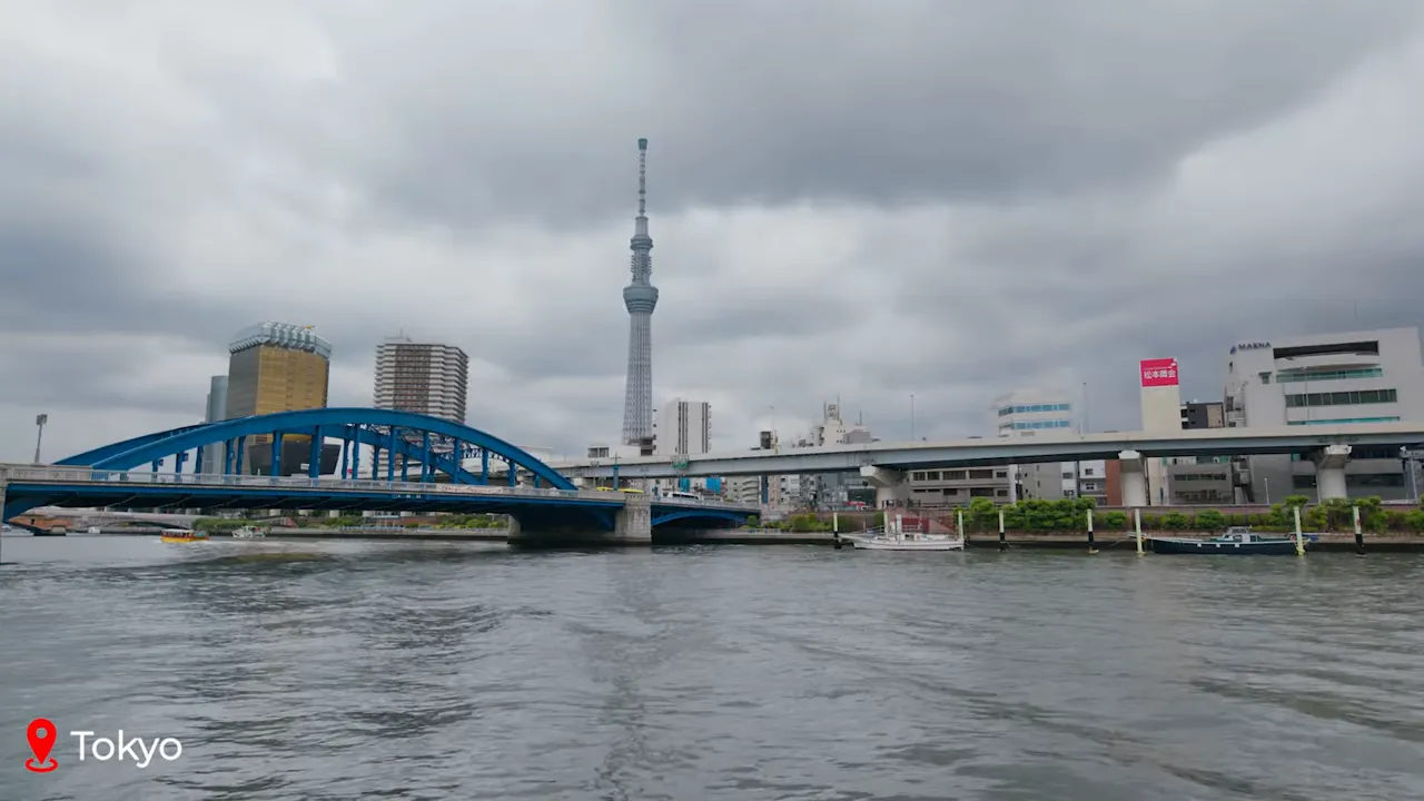 Tokyo skyline with a blue arched bridge over the Sumida River and Tokyo Skytree in the background under cloudy sky