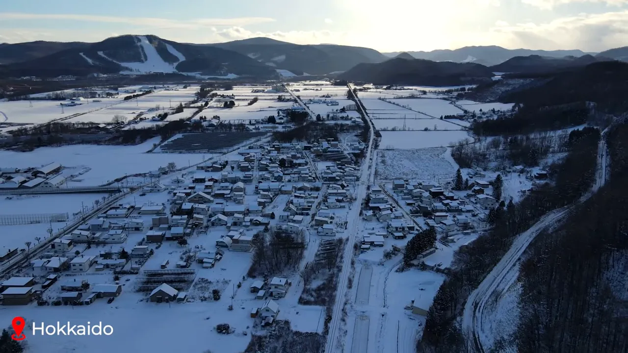 Aerial shot of a small Hokkaido village and surrounding snow‑covered fields with mountains (and ski slopes) on the horizon, labelled 'Hokkaido'.