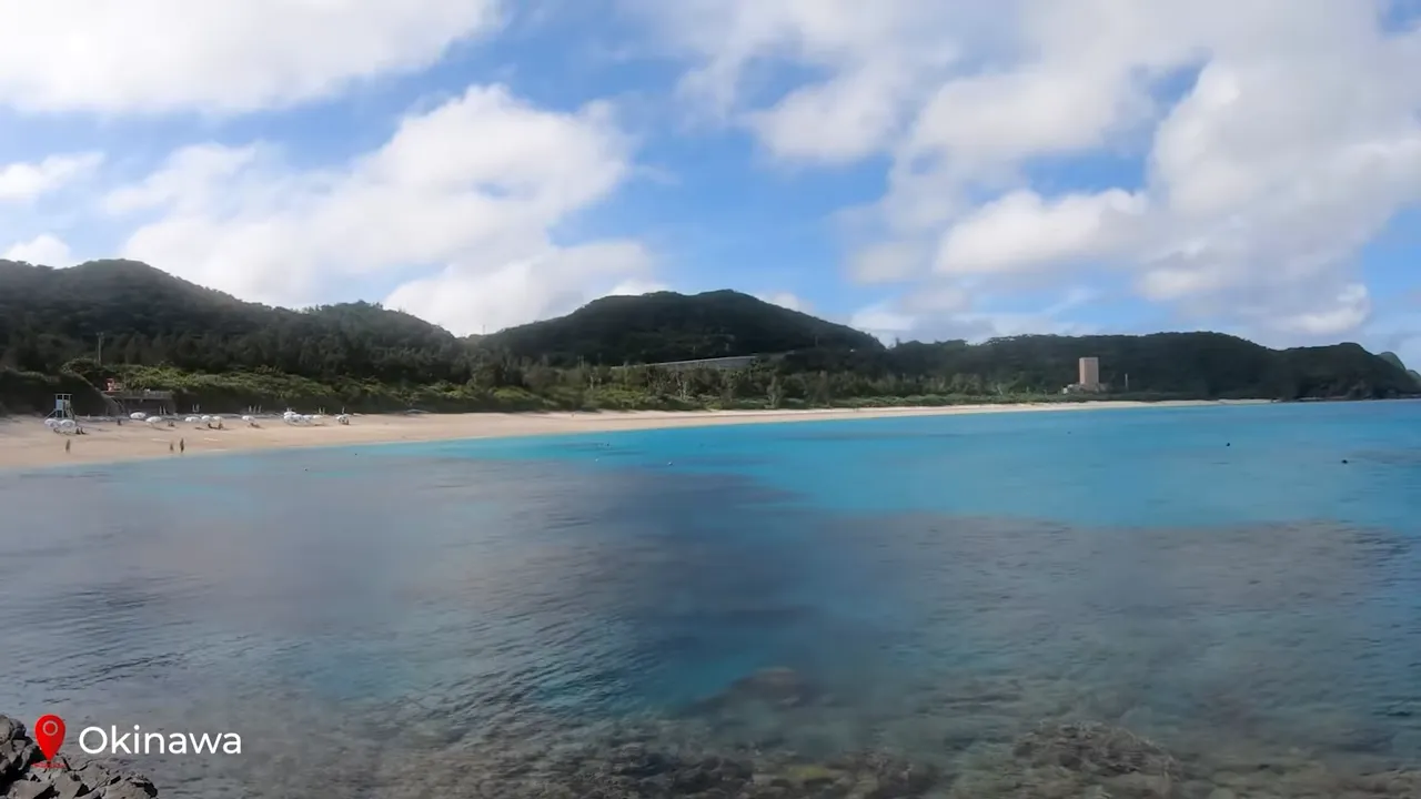 Clear turquoise water and sandy beach in Okinawa with green hills and partly cloudy sky