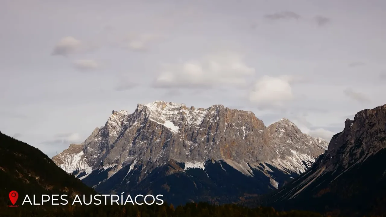 Wide panorama of the Austrian Alps with jagged, snow-dusted peaks, forested valleys below and a soft cloudy sky.