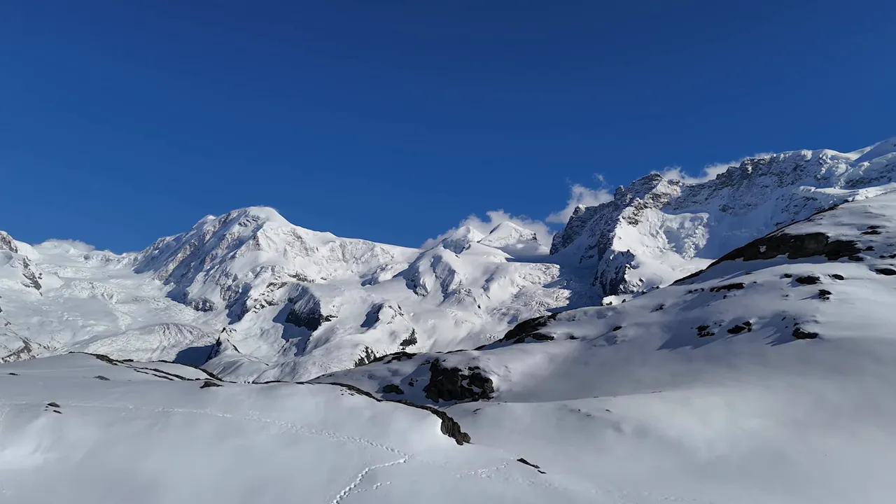 Panoramic view of the Swiss Alps with bright blue sky, snow‑covered ridges and foreground slopes showing travel tracks