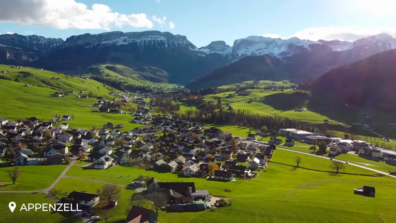 Wide aerial shot of Appenzell showing the village, expansive green fields and mountainous backdrop under clear sky