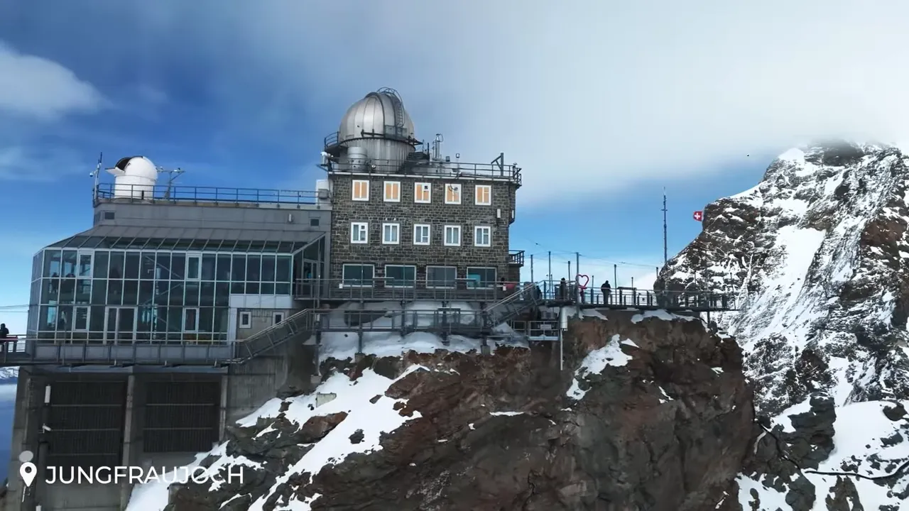 Wide clear view of the Jungfraujoch Sphinx observatory, visitor walkway and terrace perched on the cliff with snow-covered ridges behind