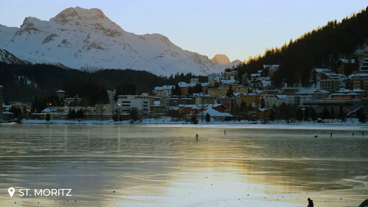 Panoramic view of frozen Lake St. Moritz reflecting warm sunset light with the lakeshore village and snow‑covered mountains behind.