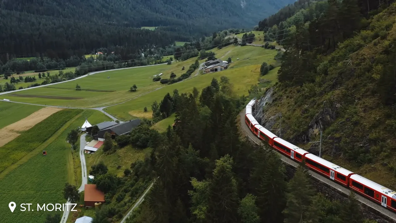 Red Glacier Express train curving on mountain tracks above a green valley with forested slopes and distant alpine fields; St. Moritz watermark.