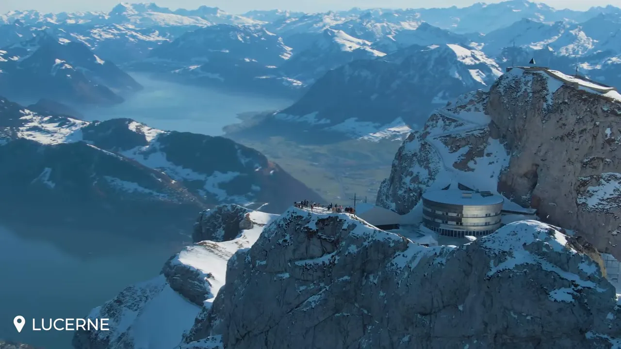 Aerial view of Mount Pilatus summit with the circular summit station, snow‑topped ridges and Lake Lucerne in the distance