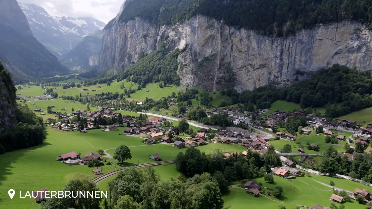 High‑clarity aerial of Lauterbrunnen showing village rooftops, curving rail line, green meadows and steep cliffs
