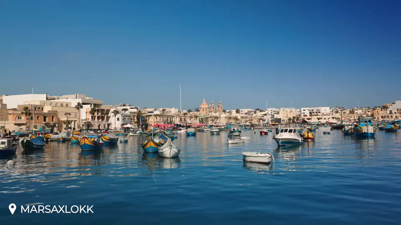 Close view of Marsaxlokk harbour with traditional colourful luzzus and small boats floating on calm, reflective water with the village skyline behind.