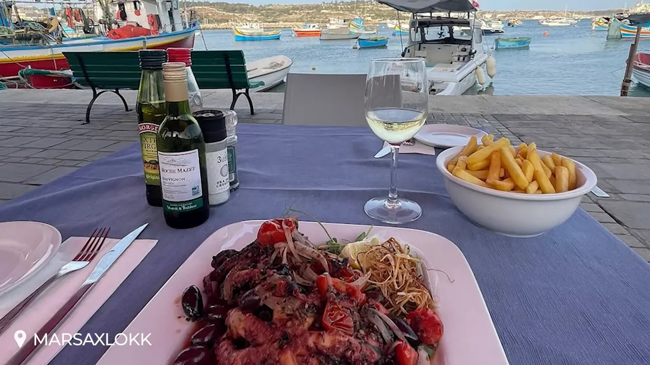 Seafood plate, wine and fries on a table with Marsaxlokk fishing boats and harbour in the background