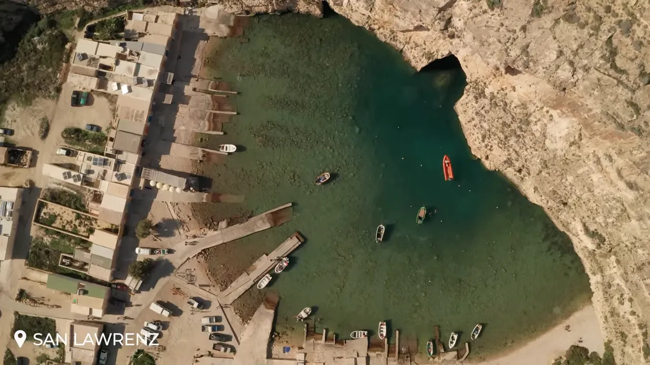 High-resolution aerial photo of San Lawrenz inlet at Dwejra with turquoise water, moored boats and a dark cave entrance in the cliff