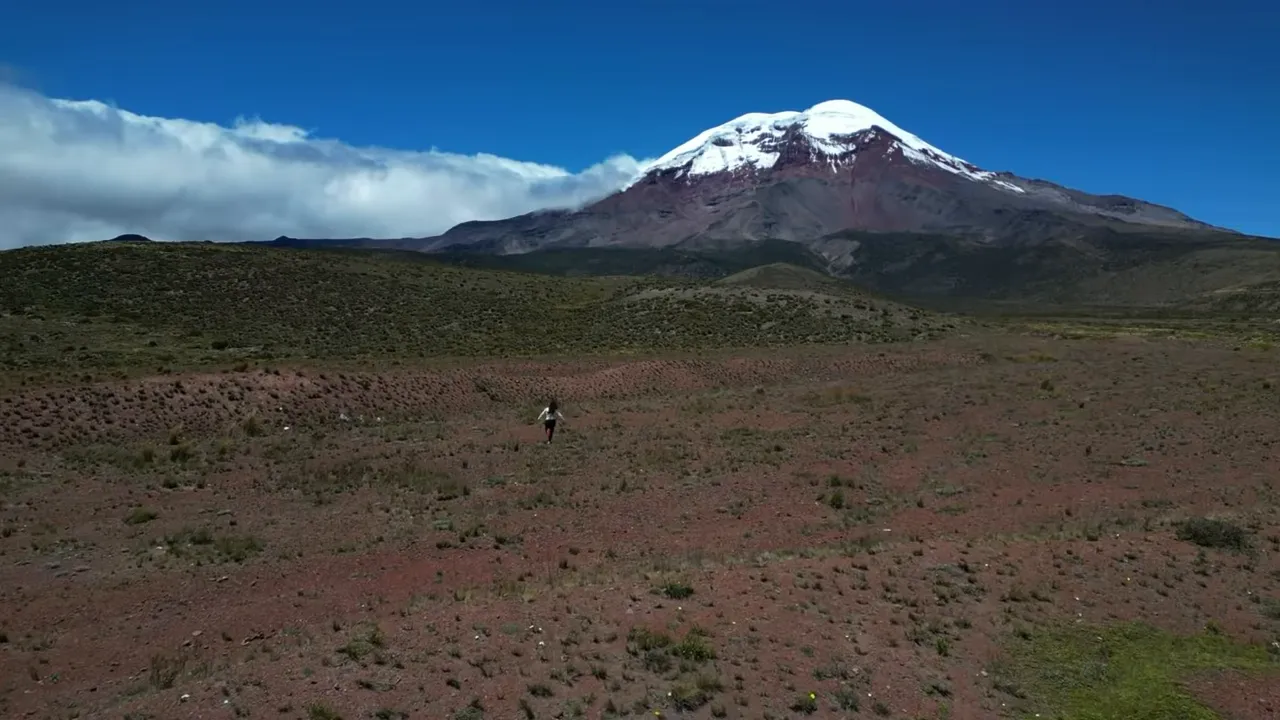 Wide landscape of Chimborazo volcano with snow-capped summit above paramo grasslands and a lone walker for scale