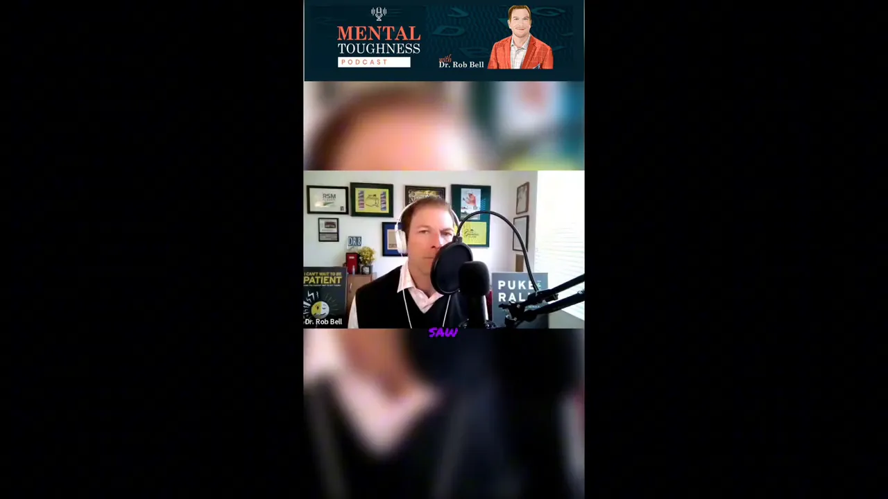 Podcast host seated in a home studio speaking into a microphone with framed awards and a sign in the background.