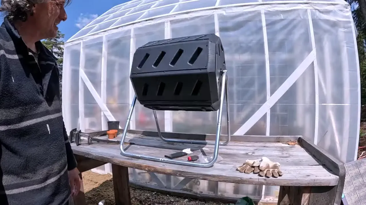 Off grid life - black compost tumbler mounted on a metal frame in front of the greenhouse