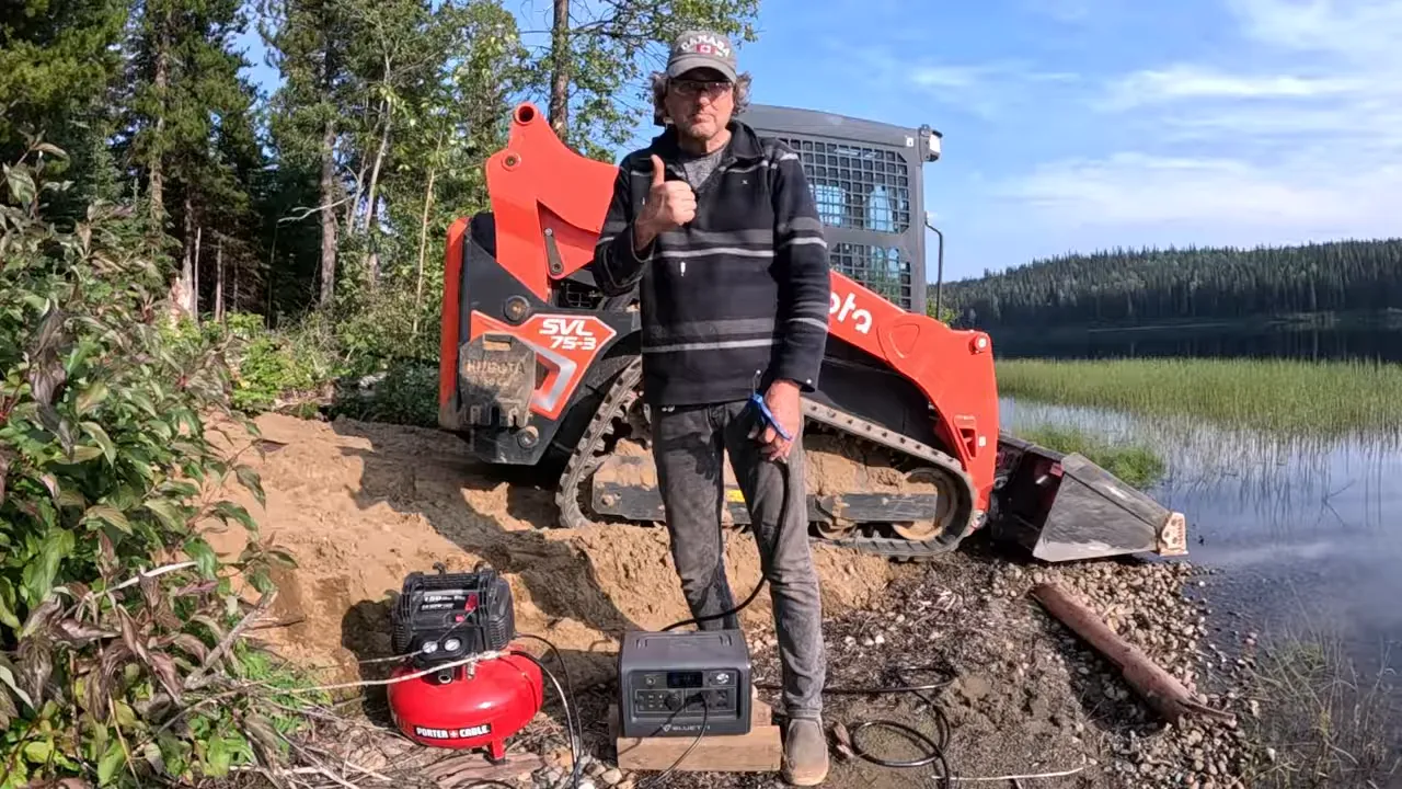 Off grid life - red compact track loader parked near the shoreline with a power station and hose nearby