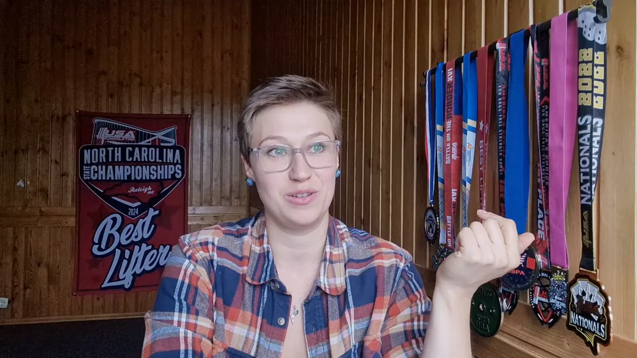 Clear head-and-shoulders shot of the presenter in a wood-paneled room with medals on the right, addressing the camera.
