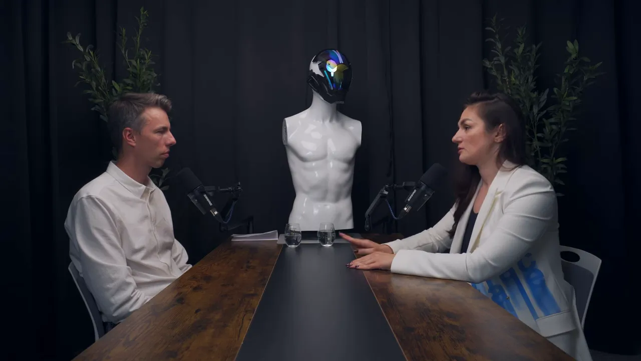 Широкий план: ведущий и гость за столом, между ними манекен с шлемом (символ AI)