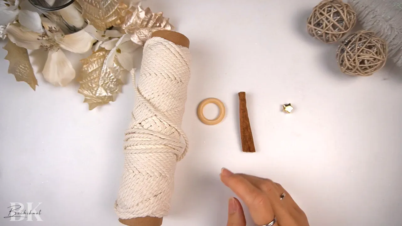 Overhead shot of a spool of macrame cord, a wooden ring, a cinnamon stick and a small gold star bead on a white background.