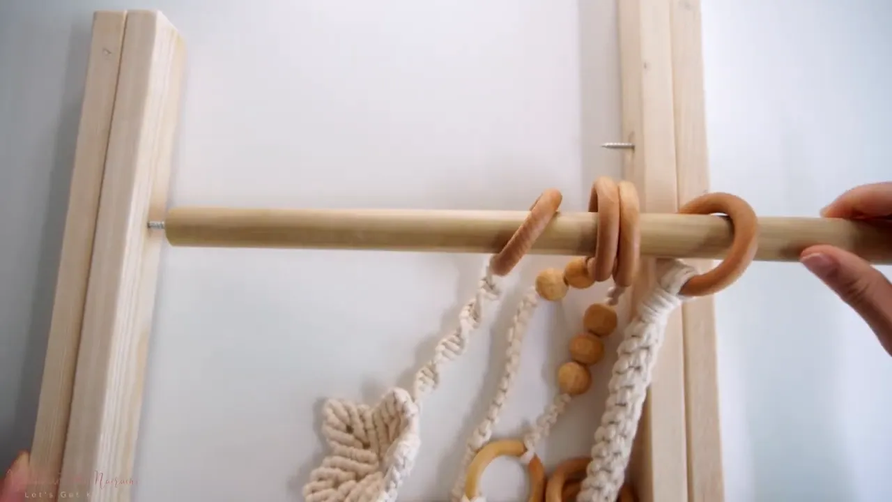 Wooden dowel with macrame toys and wooden rings slid on, held across two side frames with the left pilot screw visible — ready to be fastened.