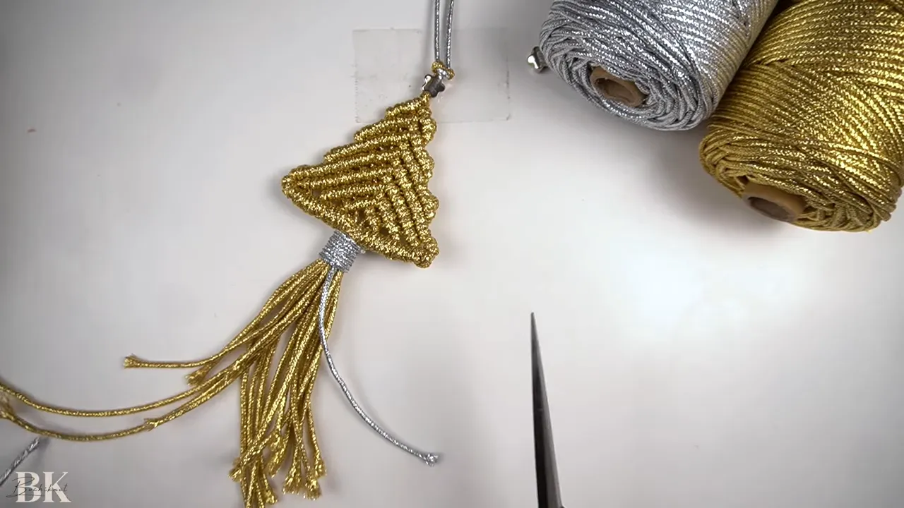 Gold macrame tree ornament laid flat with silver cord and scissors nearby on a white surface