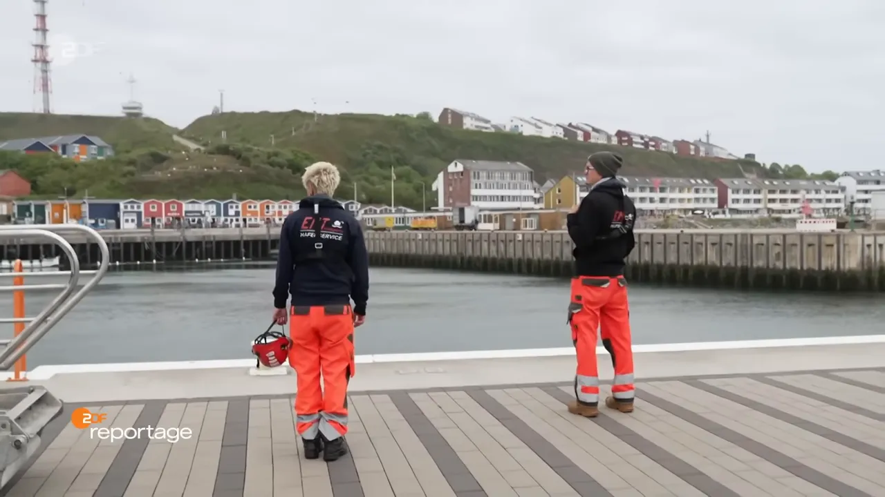 Zwei Hafenmitarbeiter in Schutzkleidung stehen auf dem Pier; im Hintergrund Helgolands bunte Häuser und das Hafenbecken.