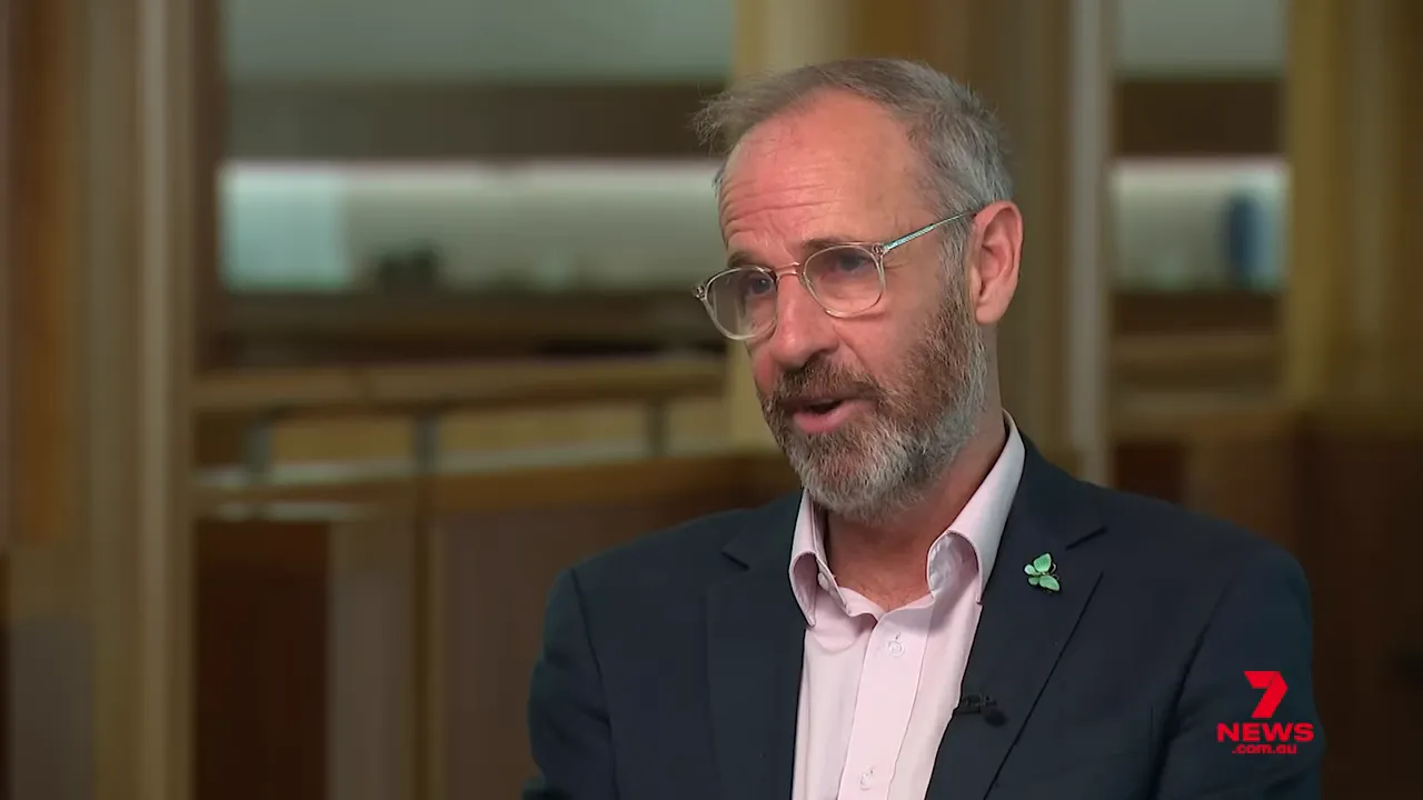 Sharp medium close-up of a bearded man in a suit with a green lapel pin speaking in an interview