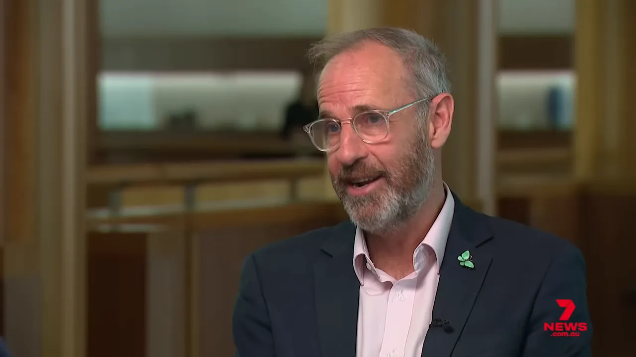 Sharp medium close-up of a Greens spokesperson in a suit with a clover lapel pin speaking in a televised interview; wood‑panel background and 7 News logo visible.