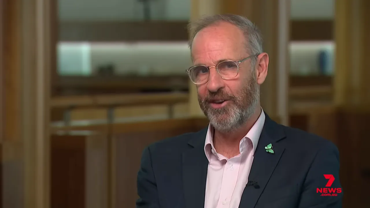 Greens spokesperson in a televised interview, clear face, engaged expression, wearing clover pin