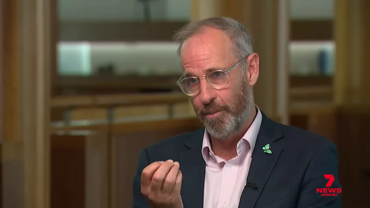 Medium close-up of a bearded man in a suit and glasses gesturing with his hand while explaining policy in an interview; 7 News watermark visible.