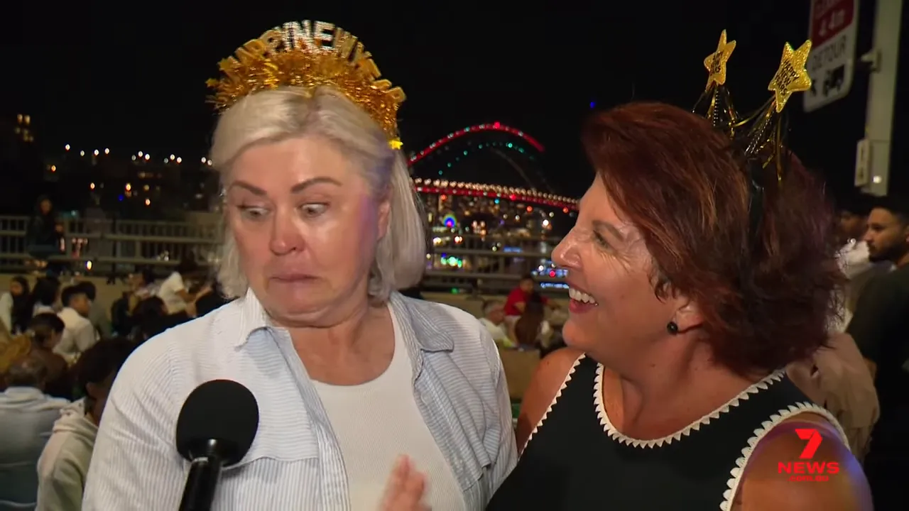 Two women wearing New Year headbands being interviewed at the harbour with the Harbour Bridge lit up in the background