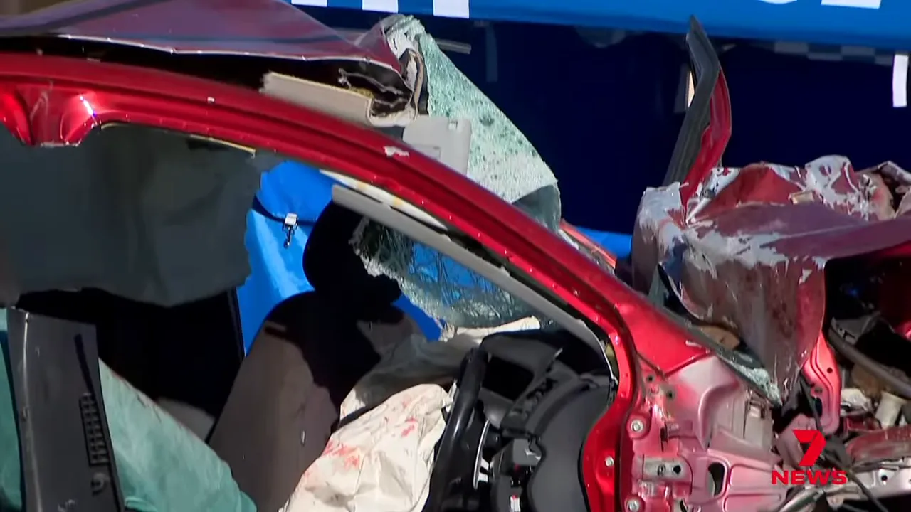 Close-up of heavily damaged red Mitsubishi showing crushed front end, shattered windshield and deployed airbags at crash scene