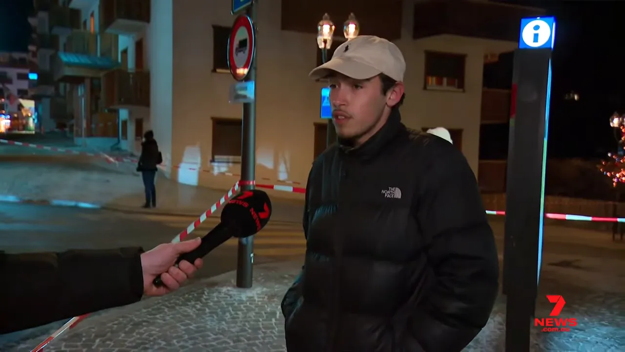 A person in a cap being interviewed beside red-and-white police tape outside a cordoned night-time scene, microphone from a news crew in frame.