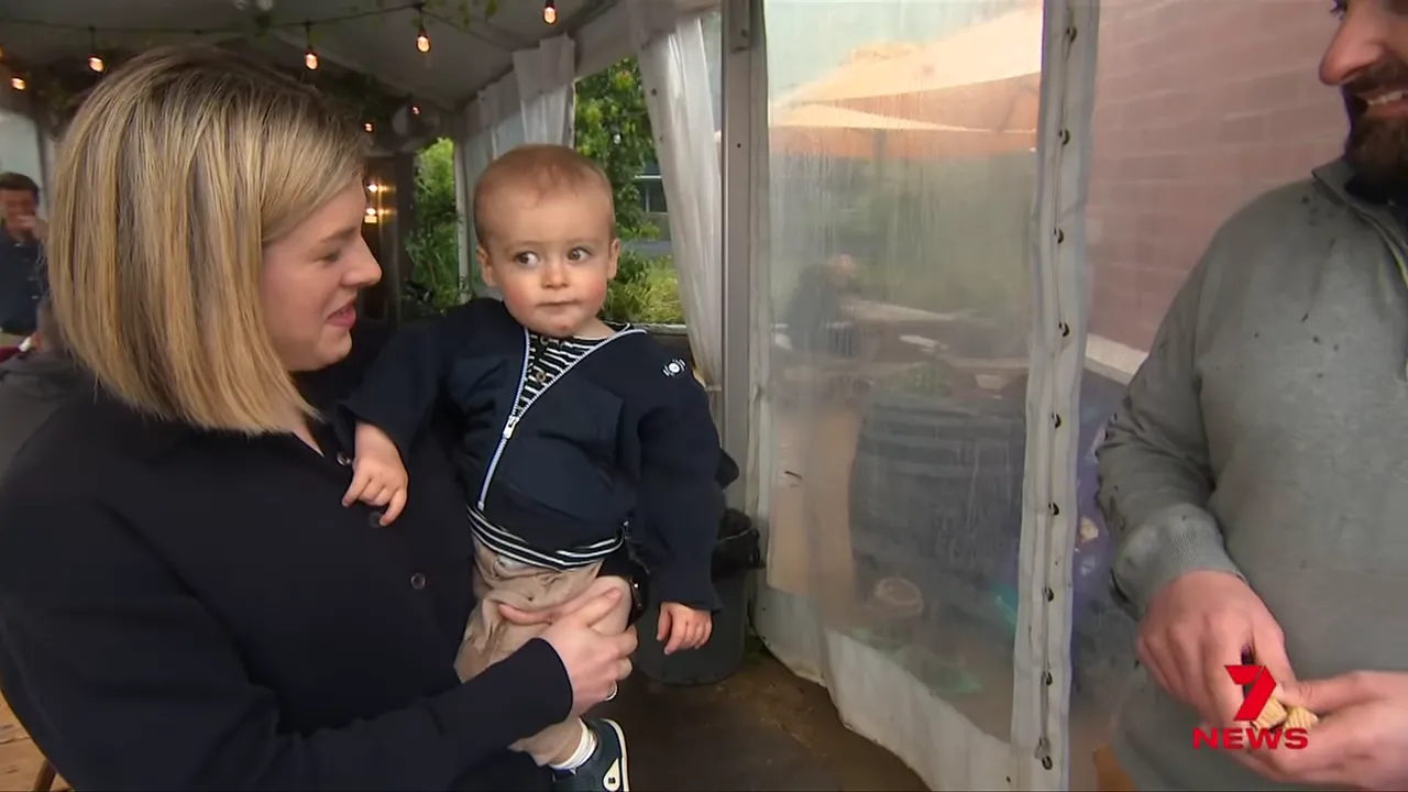 Woman holding a baby and interacting with a person off-frame inside a covered café area.
