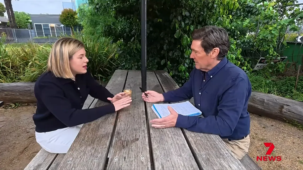Jess Wilson speaking with a male interviewer across a wooden picnic table in an outdoor setting